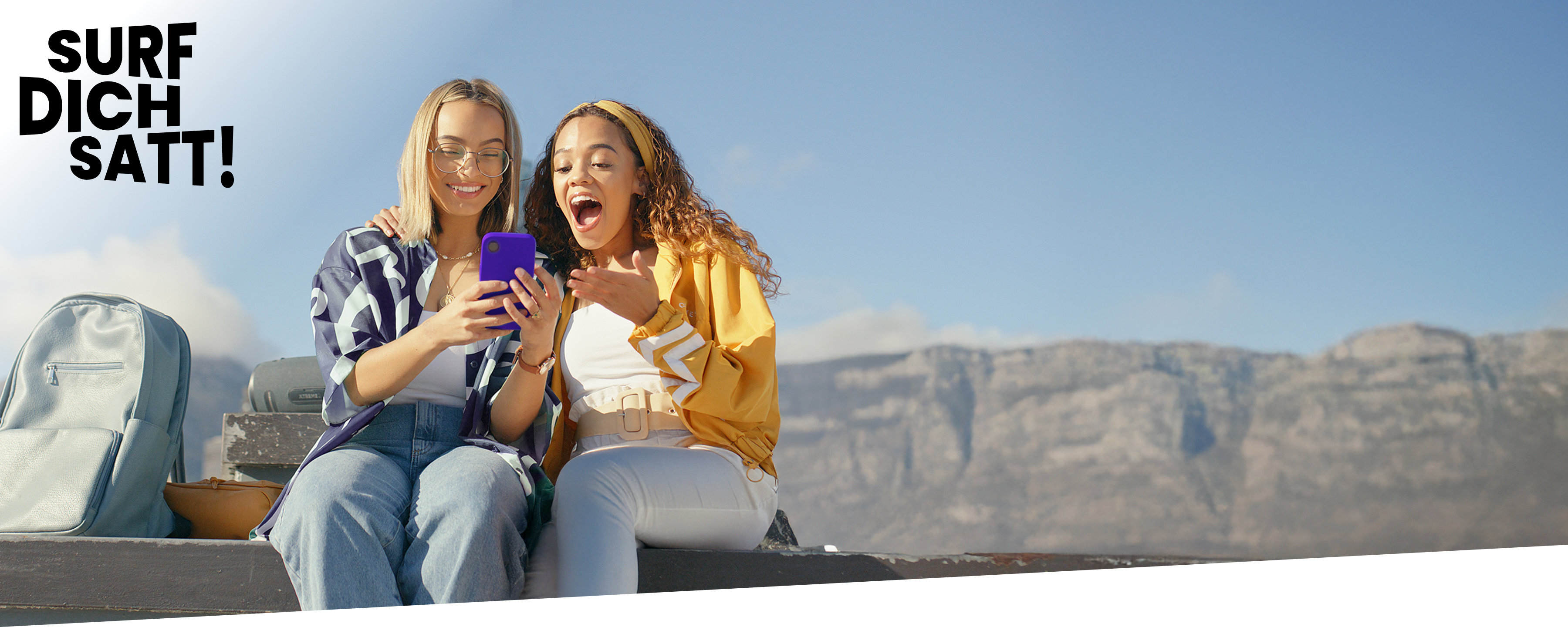 Zwei junge Frauen vor einem Bergpanorama, überrascht beim Handytarif-Vergleich auf ihrem Handy.